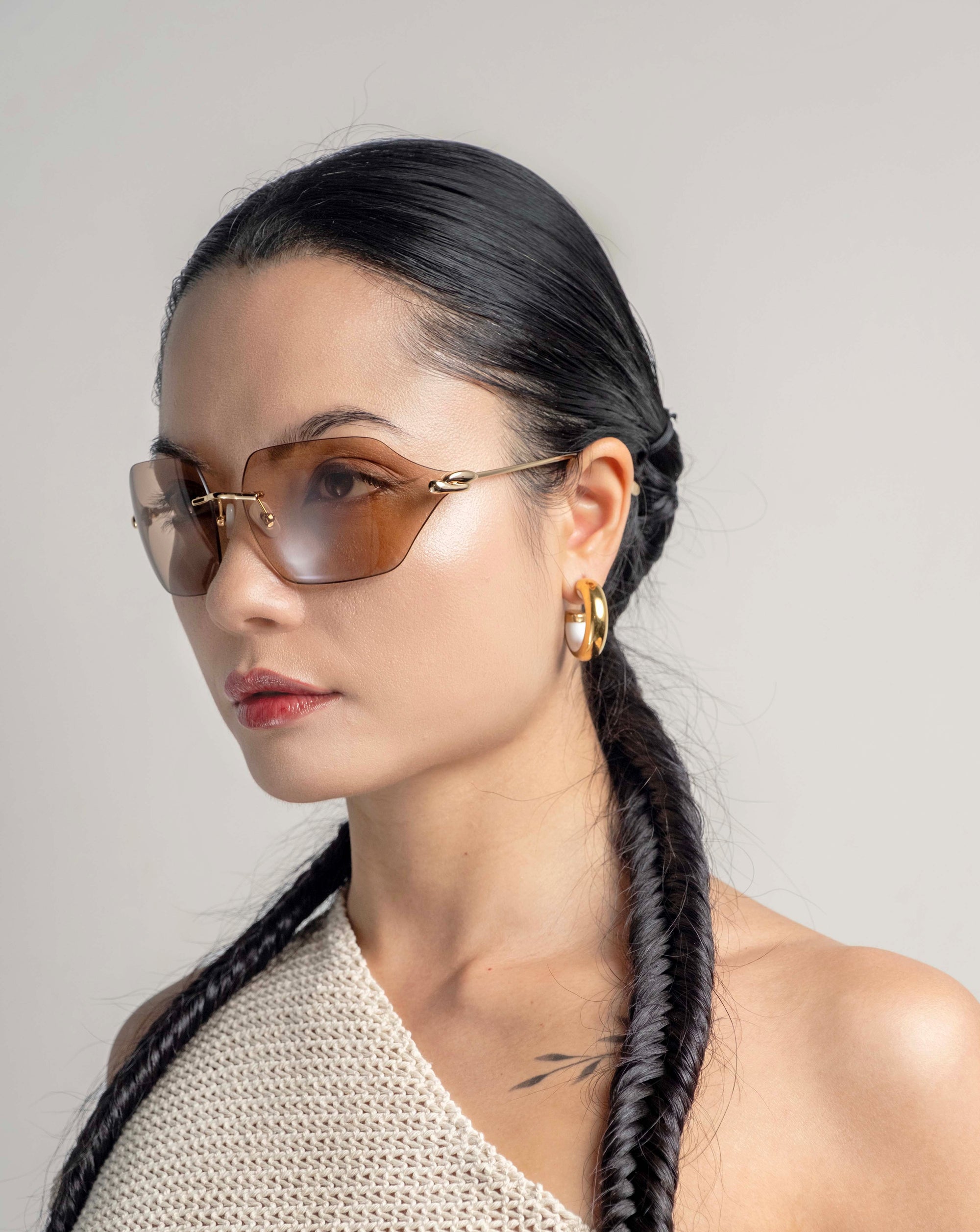 A woman with sleek dark hair fashioned into two braids, wearing For Art&#39;s Sake® Serpent II sunglasses with 18-karat gold plating and gold hoop earrings. She is dressed in a white, one-shoulder top and is gazing to the side against a plain background. A small tattoo is visible on her shoulder.