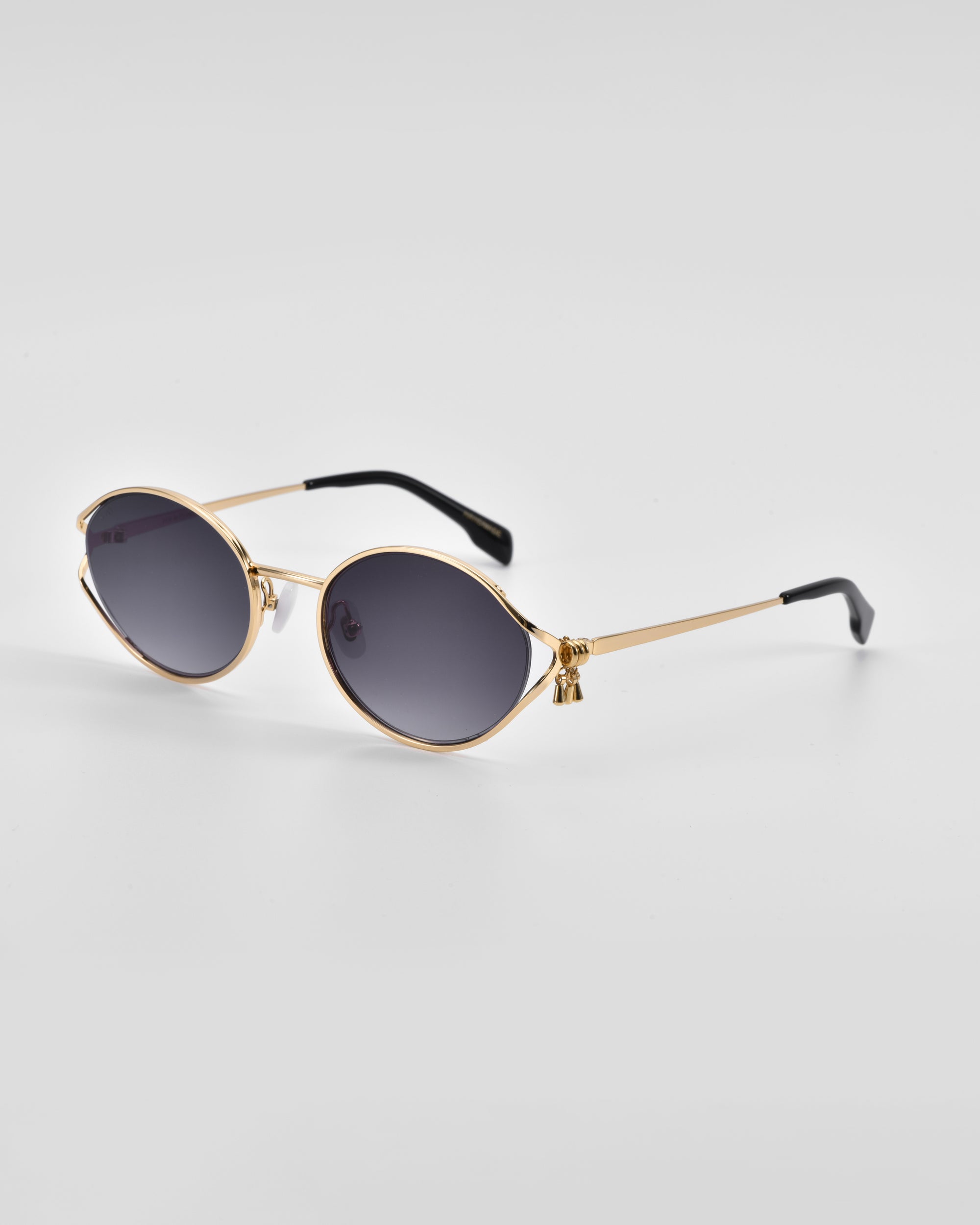 A pair of stylish For Art&#39;s Sake® Sky sunglasses with golden metal frames and dark, oval lenses. The temples have black tips, providing a contrast against the 18-karat gold. The natural jade-stone nose pads add an extra touch of luxury. The background is a clean white surface, highlighting the elegant design of the sunglasses.