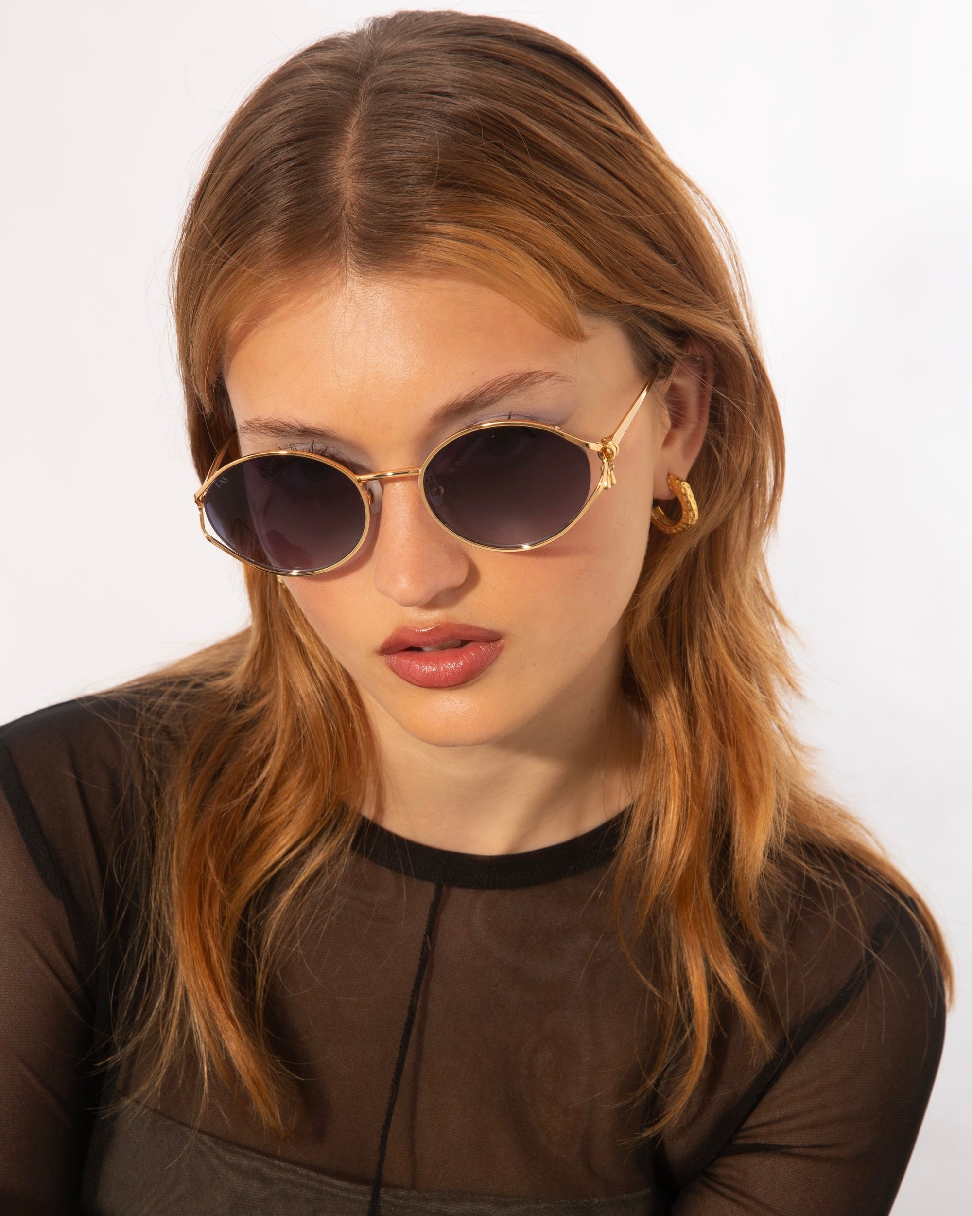 A person with long, light brown hair is wearing round Sky sunglasses by For Art's Sake® featuring jade-stone nose pads and 18-karat gold plating, along with small gold hoop earrings. They are dressed in a black, semi-sheer top and looking slightly downward against a plain white background.