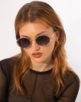 A person with long, light brown hair is wearing round Sky sunglasses by For Art's Sake® featuring jade-stone nose pads and 18-karat gold plating, along with small gold hoop earrings. They are dressed in a black, semi-sheer top and looking slightly downward against a plain white background.