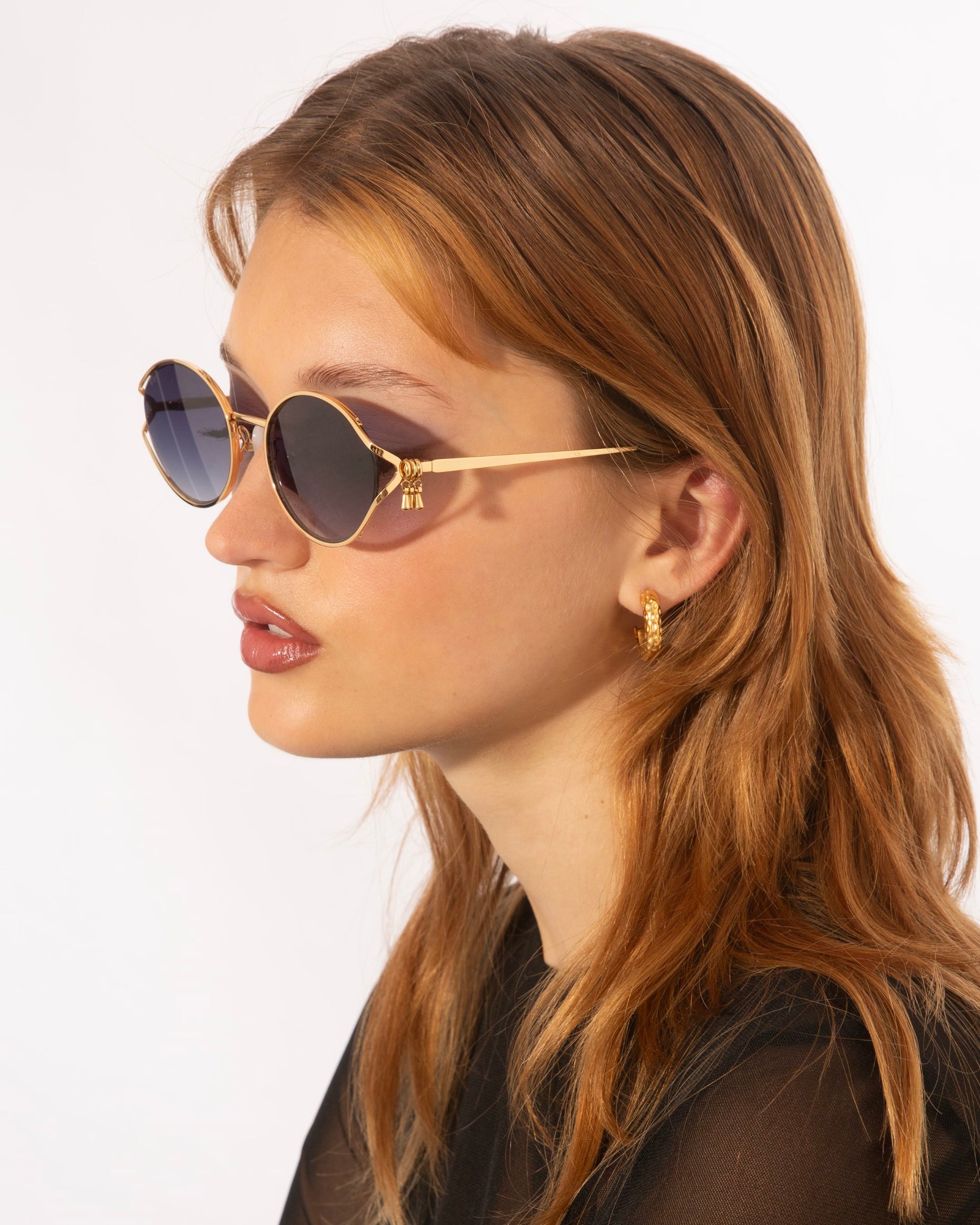 A young person with long, light brown hair is wearing round, dark For Art's Sake® Sky sunglasses adorned with jade-stone nose pads and small hoop earrings with 18-karat gold plating. They are looking off to the side, and their lips are slightly parted. The background is plain white.
