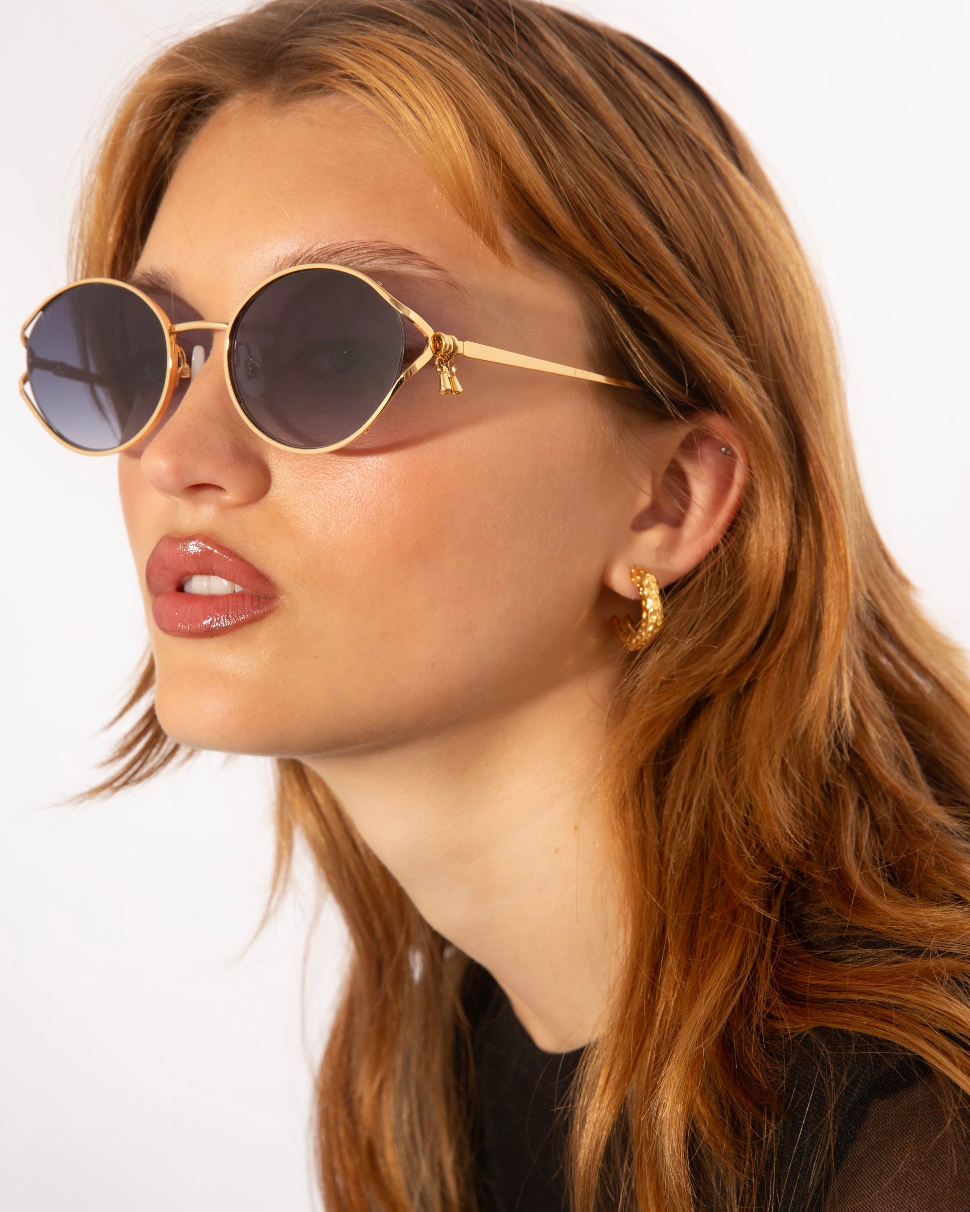 A person with long, wavy, light brown hair is wearing round, gold-rimmed sunglasses from For Art&#39;s Sake® featuring jade-stone nose pads and gold hoop earrings. They are looking slightly to the side with a neutral expression. The background is plain white. The sunglasses are called Sky.