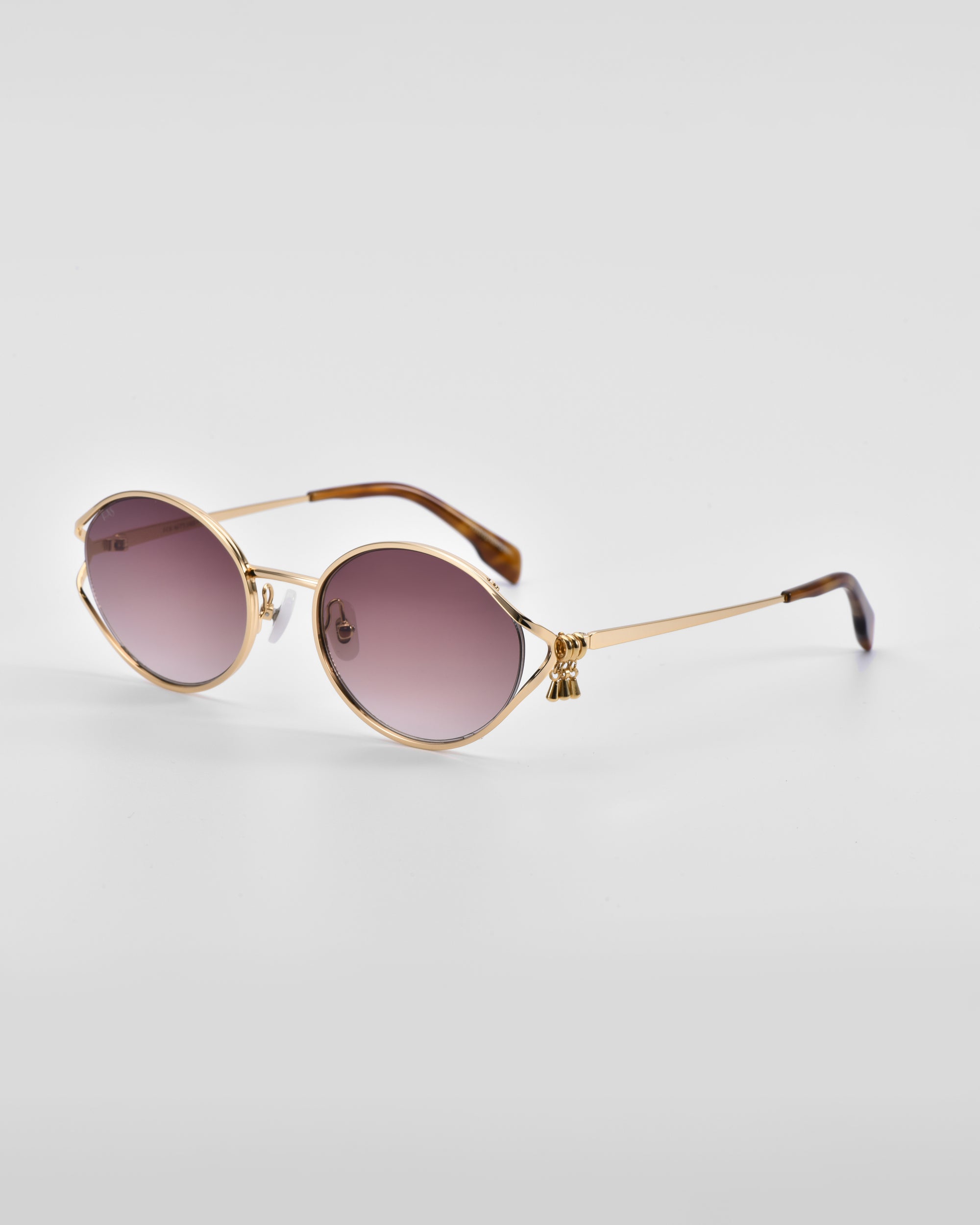 A pair of stylish &#39;Sky&#39; sunglasses by For Art&#39;s Sake® with 18-karat gold frames and oval, pink-tinted lenses. The design includes slender arms with black-tipped ends and small decorative tassels hanging from the temples, as well as natural jade-stone nose pads. Placed against a plain white background.