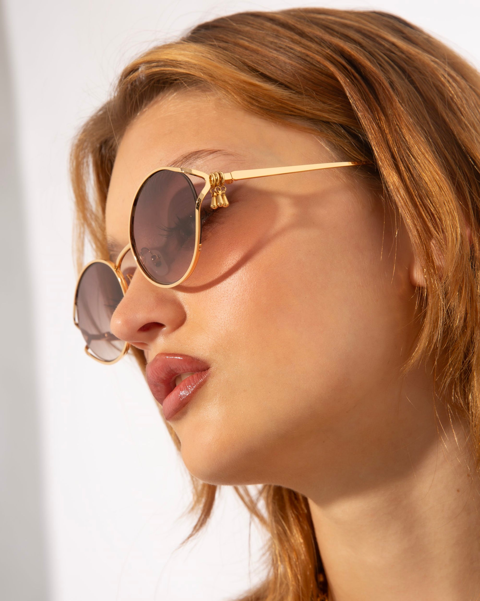 A person with light brown hair wearing round, 18-karat gold-framed Sky sunglasses by For Art&#39;s Sake® looks off to the side. The background is bright and minimalist, highlighting the person&#39;s face and stylish eyewear.