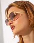 A person with light brown hair wearing round, 18-karat gold-framed Sky sunglasses by For Art's Sake® looks off to the side. The background is bright and minimalist, highlighting the person's face and stylish eyewear.