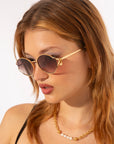 A person with long, light-brown hair is wearing round, black Sky sunglasses with gold accents and a gold and pearl necklace. Featuring jade-stone nose pads, the stylish For Art's Sake® Sky sunglasses add an extra touch of luxury as they look down and to the left against a white background.