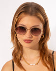 A young woman with auburn hair wears round For Art's Sake® Sky sunglasses featuring gold frames and jade-stone nose pads, complemented by 18-karat gold hoop earrings and a gold necklace with pearl accents. She has a confident expression and is set against a plain white background.