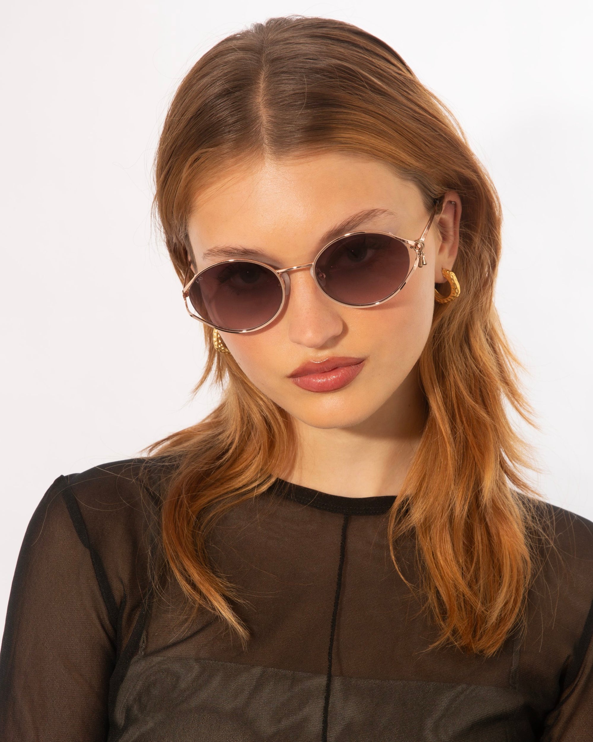 A person with long, light brown hair is wearing round, dark Sky sunglasses by For Art&#39;s Sake® with jade-stone nose pads and gold hoop earrings. They have a neutral expression and are dressed in a sheer black top, posing against a plain white background.