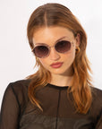 A person with long, light brown hair is wearing round, dark Sky sunglasses by For Art's Sake® with jade-stone nose pads and gold hoop earrings. They have a neutral expression and are dressed in a sheer black top, posing against a plain white background.