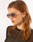 A person with long, light brown hair is wearing round *For Art's Sake® Sky* sunglasses featuring jade-stone nose pads and gold hoop earrings. They are dressed in a black sheer top and are turned slightly to the side with a neutral expression. The background is plain white.