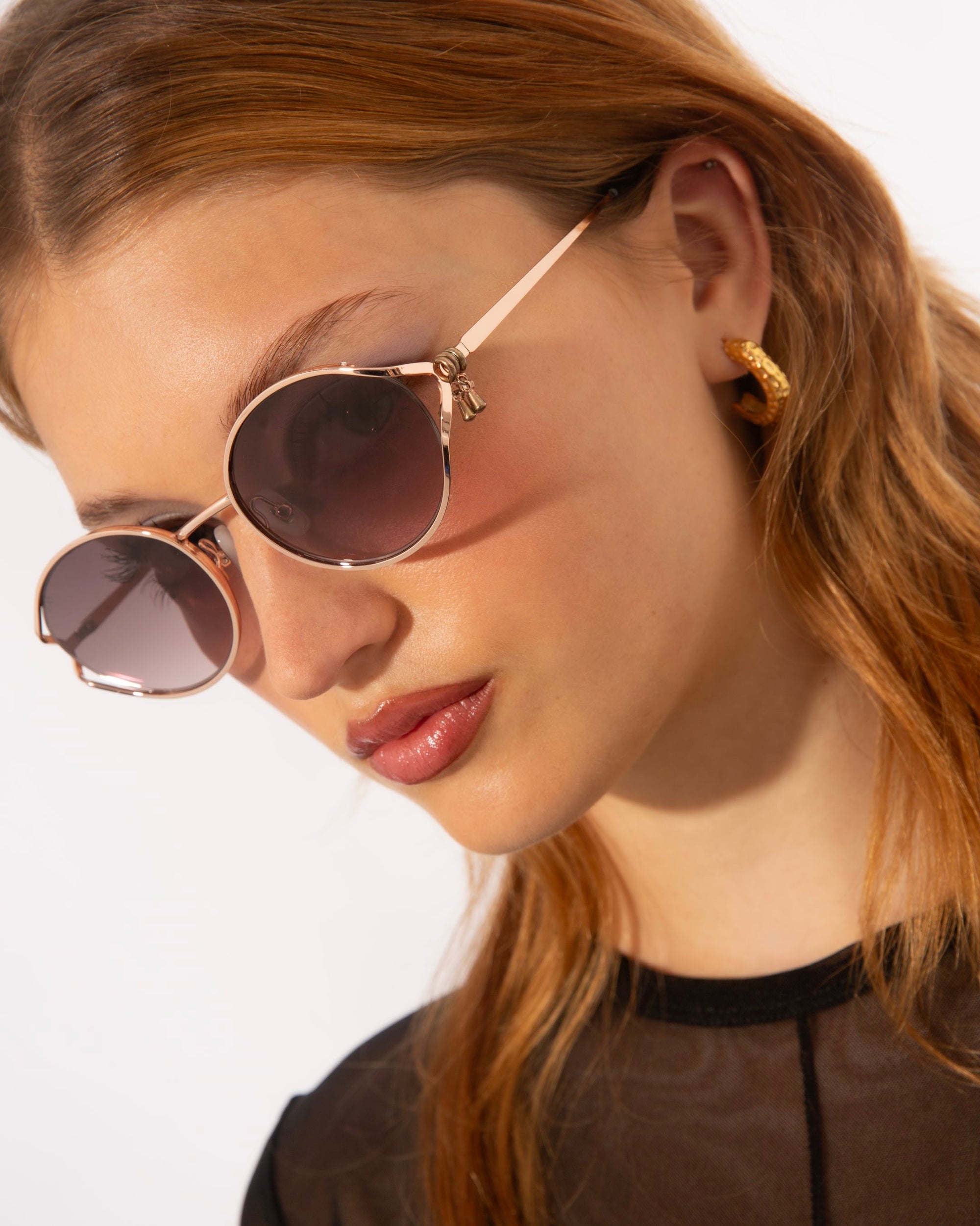 A person with long, light brown hair is wearing For Art&#39;s Sake® Sky sunglasses with jade-stone nose pads and gold hoop earrings. They are looking slightly downwards with a neutral expression while sporting a black, semi-sheer top. The background is plain white.