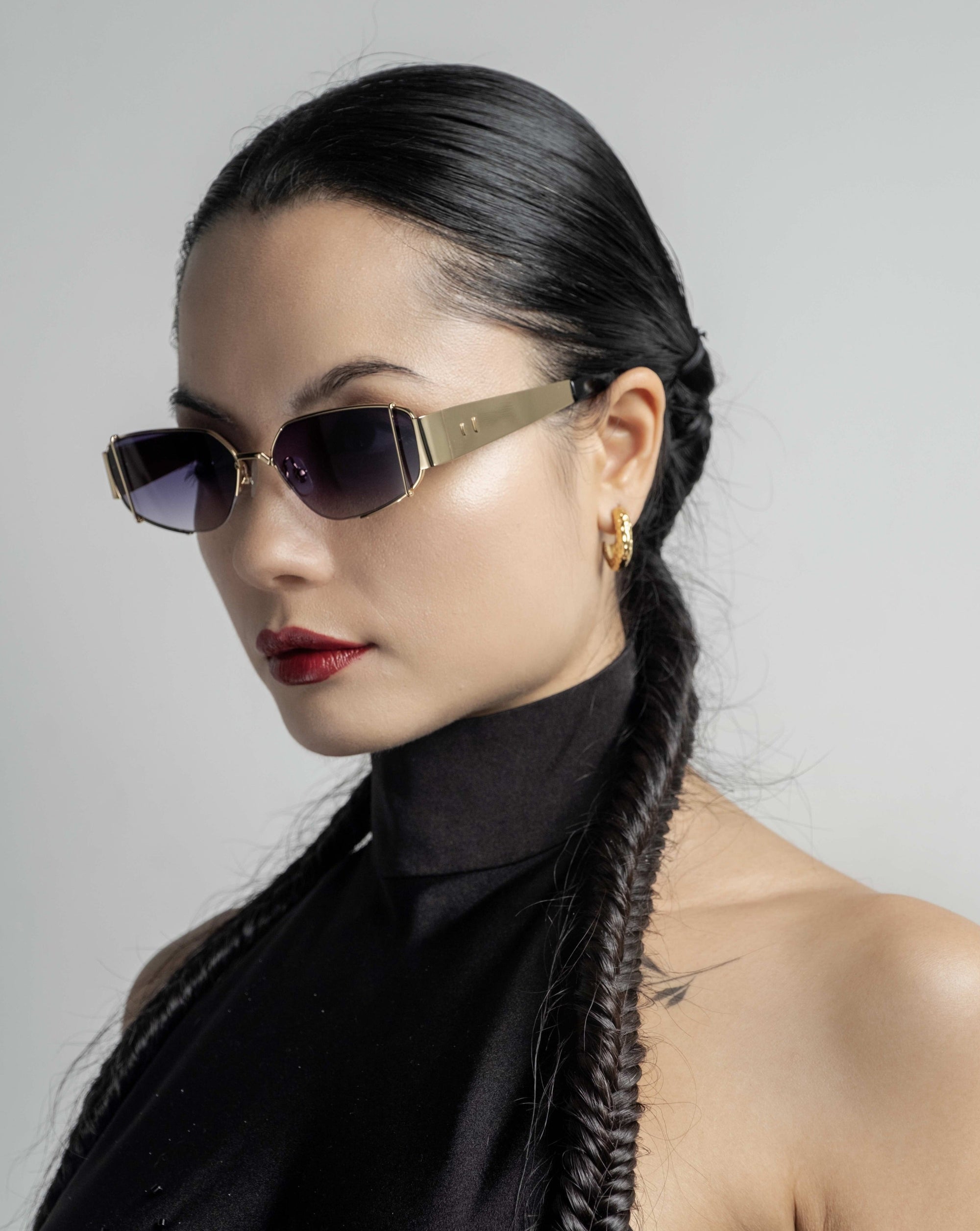 A woman with sleek black hair in braids, wearing Talia sunglasses by For Art&#39;s Sake® with metal frames and a black sleeveless high-collar top. She has 18-karat gold hoop earrings and a small tattoo on her shoulder. The background is a plain light gray.