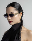 A woman with sleek black hair in braids, wearing Talia sunglasses by For Art's Sake® with metal frames and a black sleeveless high-collar top. She has 18-karat gold hoop earrings and a small tattoo on her shoulder. The background is a plain light gray.