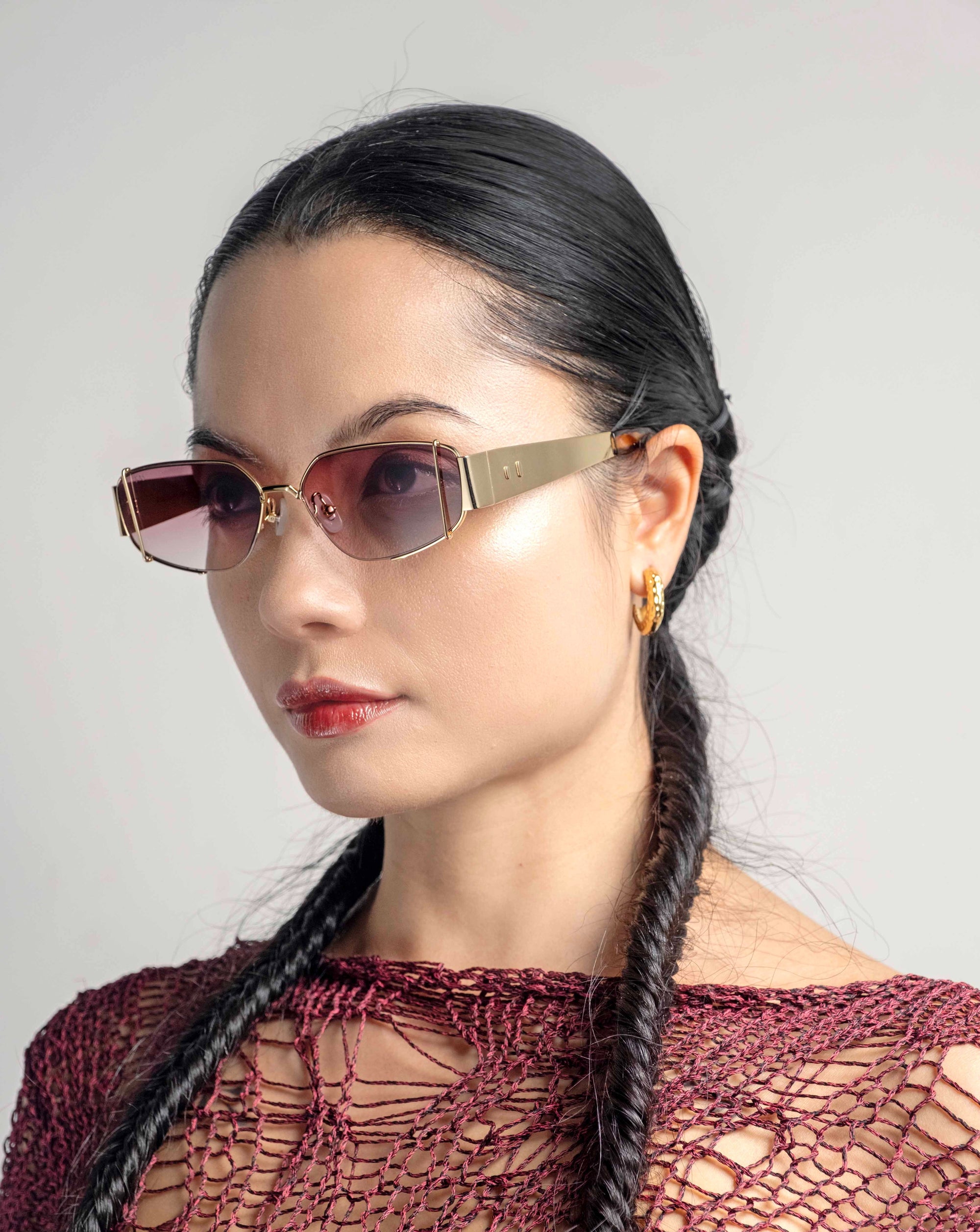 A woman with long, dark braided hair is wearing stylish For Art&#39;s Sake® Talia sunglasses with 18-karat gold metal frames, gold hoop earrings, and a red, intricately knitted top. She is looking to the side against a plain, light-colored background.