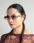 A woman with long, dark braided hair is wearing stylish For Art's Sake® Talia sunglasses with 18-karat gold metal frames, gold hoop earrings, and a red, intricately knitted top. She is looking to the side against a plain, light-colored background.