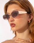 A person with light hair is wearing stylish, angular Talia sunglasses from For Art's Sake®, featuring dark lenses and gold metal frames with jade-stone nose pads. They have gold hoop earrings and glossy lip makeup. The background is plain white, and the individual gazes slightly to the side.