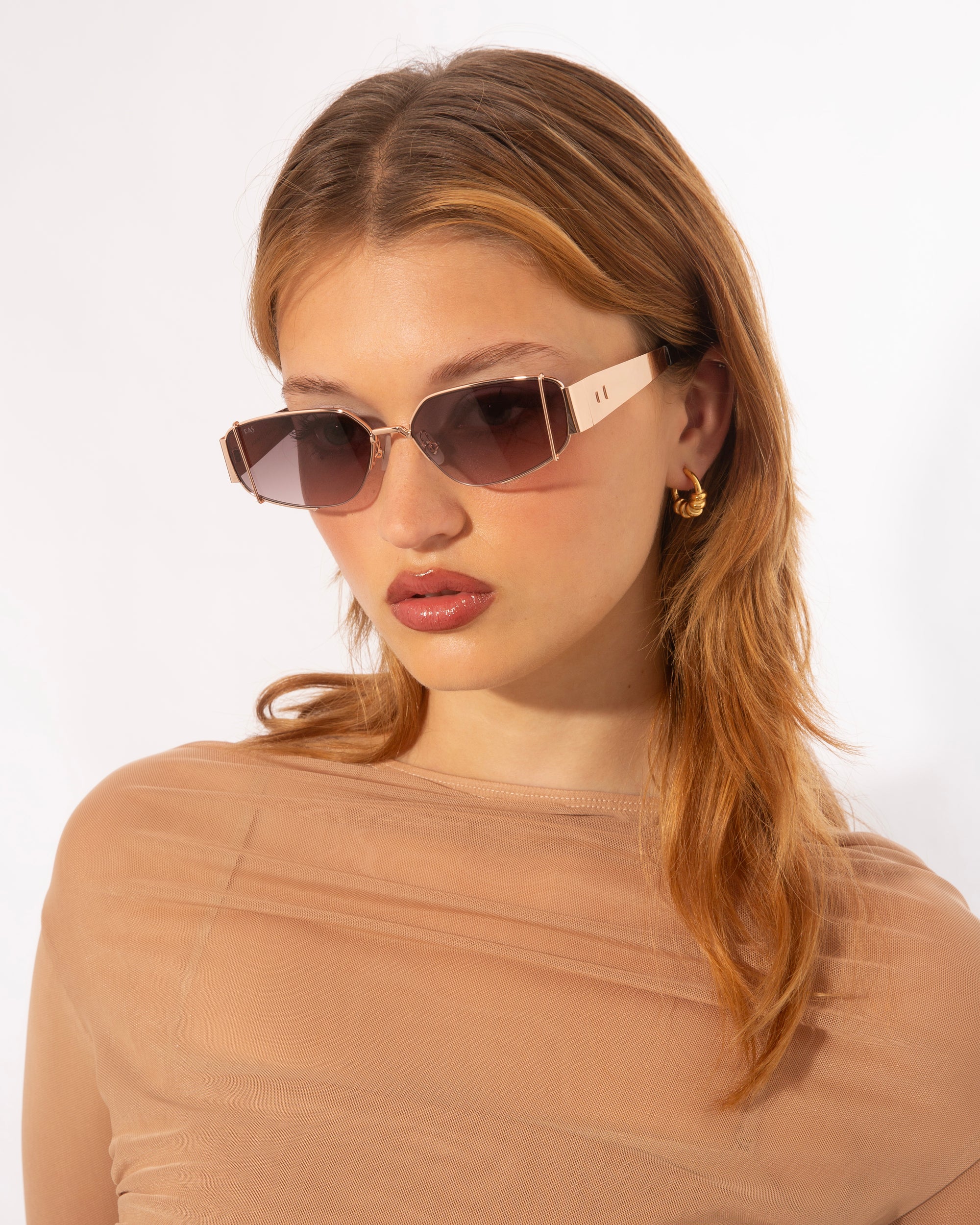 A person with long, light brown hair is wearing rectangular, gradient-lens sunglasses with full metal frames from For Art&#39;s Sake® called Talia. They are also wearing a sheer, light brown top and 18-karat gold hoop earrings. The background is plain white.