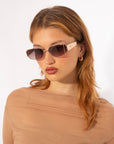 A person with long, light brown hair is wearing rectangular, gradient-lens sunglasses with full metal frames from For Art's Sake® called Talia. They are also wearing a sheer, light brown top and 18-karat gold hoop earrings. The background is plain white.