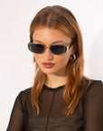 A person with long, straight, light brown hair is wearing Talia sunglasses by For Art's Sake® with metal frames, 18-karat gold hoop earrings, and a sheer black top. They are gazing slightly downward against a plain white background.