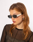 A person with light brown hair wearing rectangular Talia sunglasses by For Art's Sake® featuring jade-stone nose pads and silver hoop earrings is pictured against a white background. They are dressed in a black, semi-sheer top, looking slightly to the right.