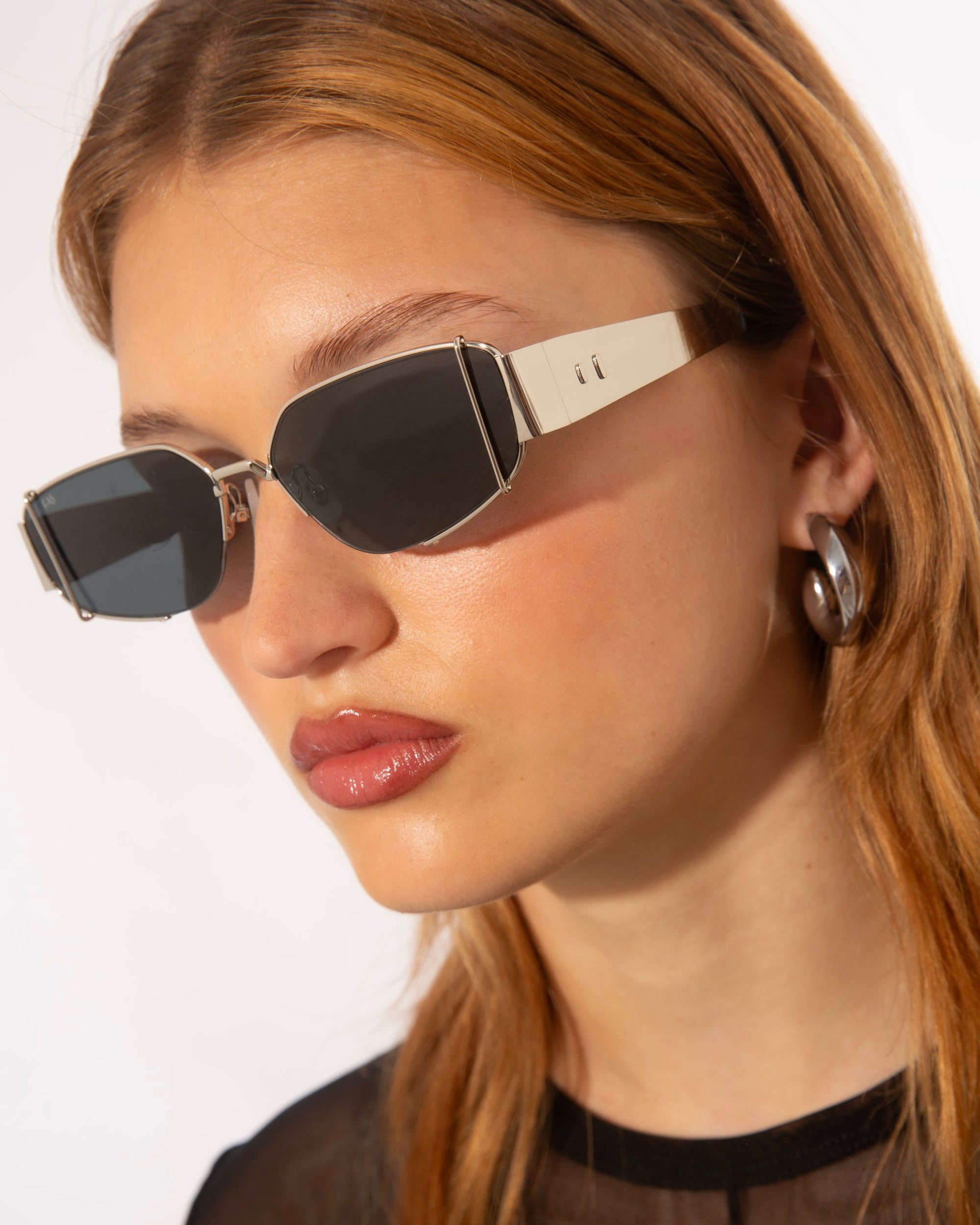Close-up of a person with long light brown hair wearing modern angular Talia sunglasses from For Art&#39;s Sake® featuring thick white frames. They have neutral makeup and are also wearing large, silver hoop earrings. The background is plain and white, highlighting the accessories.