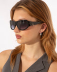 A woman with light brown hair wearing oversized For Art's Sake® Raven sunglasses and a sleeveless gray top. She has silver drop earrings and is gazing to the side against a plain white background.