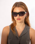 A person with long, light brown hair is wearing large, Y2K wrap sunglasses and silver hoop earrings. They are dressed in a sleeveless gray blazer and are posing against a plain white background, showing off the Raven by For Art's Sake®.