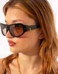 A person with light brown hair wearing For Art's Sake® Raven wrap sunglasses with tinted lenses looks to the side. They are also adorned with an 18-karat gold chain necklace. The background is plain white.