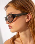 A person with light brown hair is wearing Raven sunglasses by For Art's Sake® and an 18-karat gold chain necklace. The person is facing sideways to the right, displaying glossy, natural-looking lips, and wearing a small earring. The background is a plain, light-colored wall.