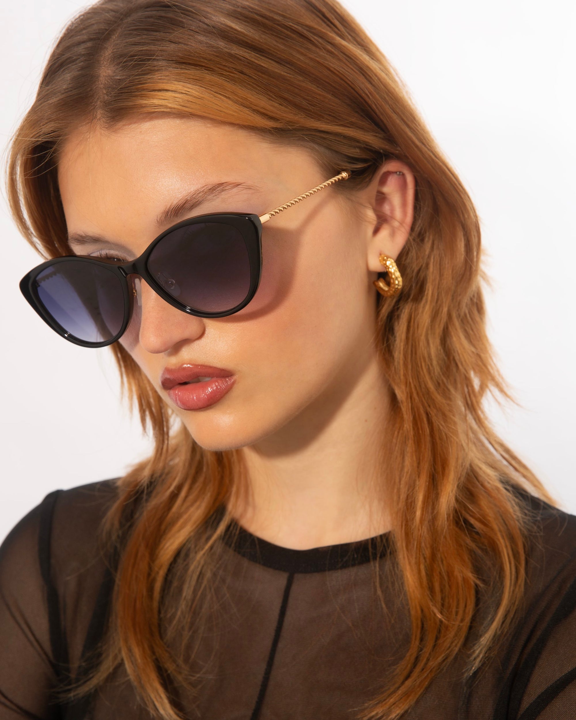 A person with long, auburn hair and wearing a black sheer top and black cat-eye sunglasses featuring jade-stone nose pads by For Art's Sake® Perla II is looking down. They have gold hoop earrings and a slightly serious expression. The background is plain white.
