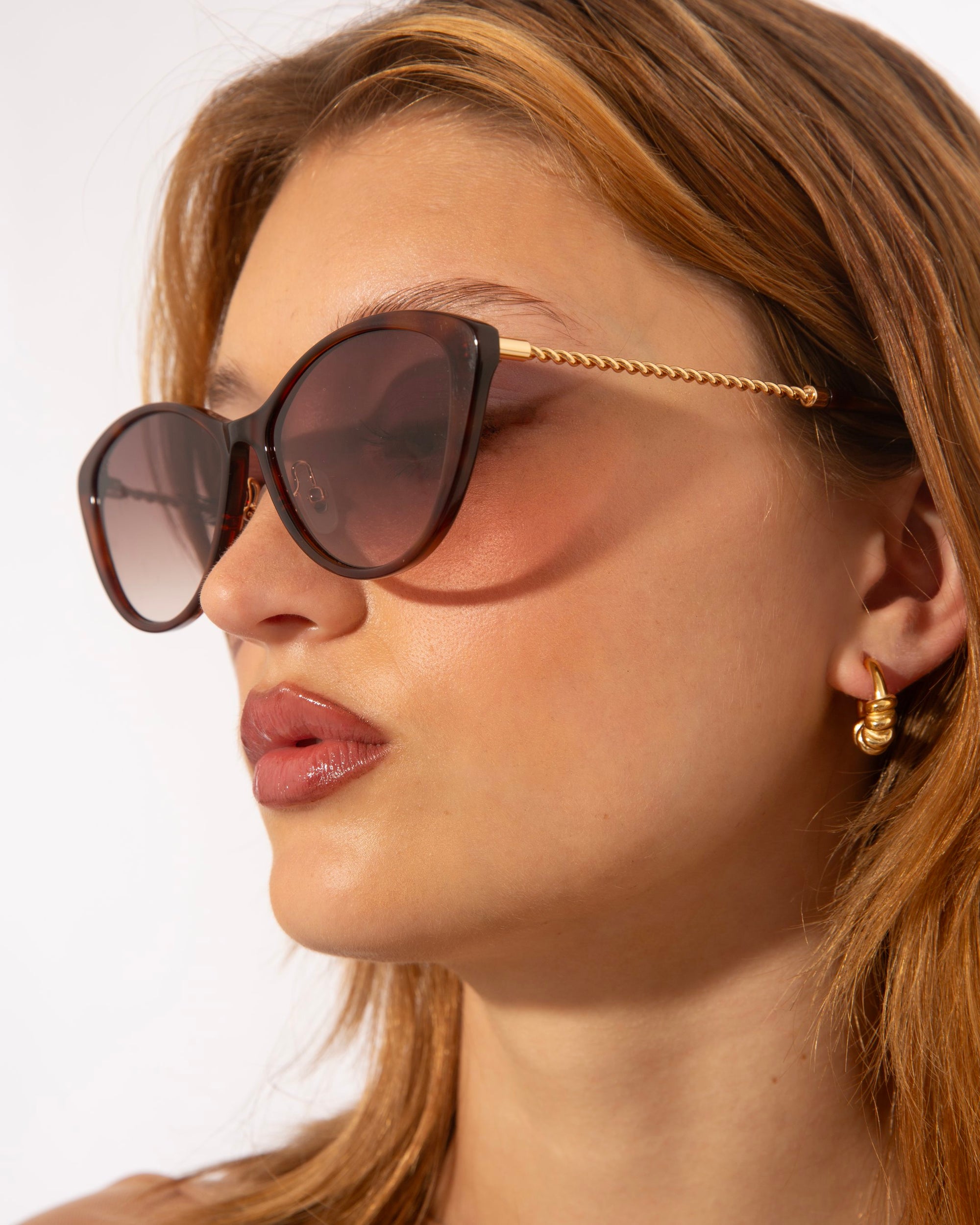 A close-up of a person wearing large, dark, cat-eye For Art&#39;s Sake® Perla II sunglasses with a gold frame and jade-stone nose pads. They have light brown hair and are also wearing a small 18-karat gold hoop earring in one ear. The background is plain white.