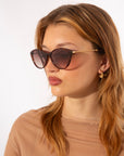 A woman with light brown hair is wearing large, dark-tinted For Art's Sake® Perla II sunglasses with a braided detail on the arms and 18-karat gold accents. She has small gold hoop earrings and is dressed in a tan, sheer top. The background is a plain white.