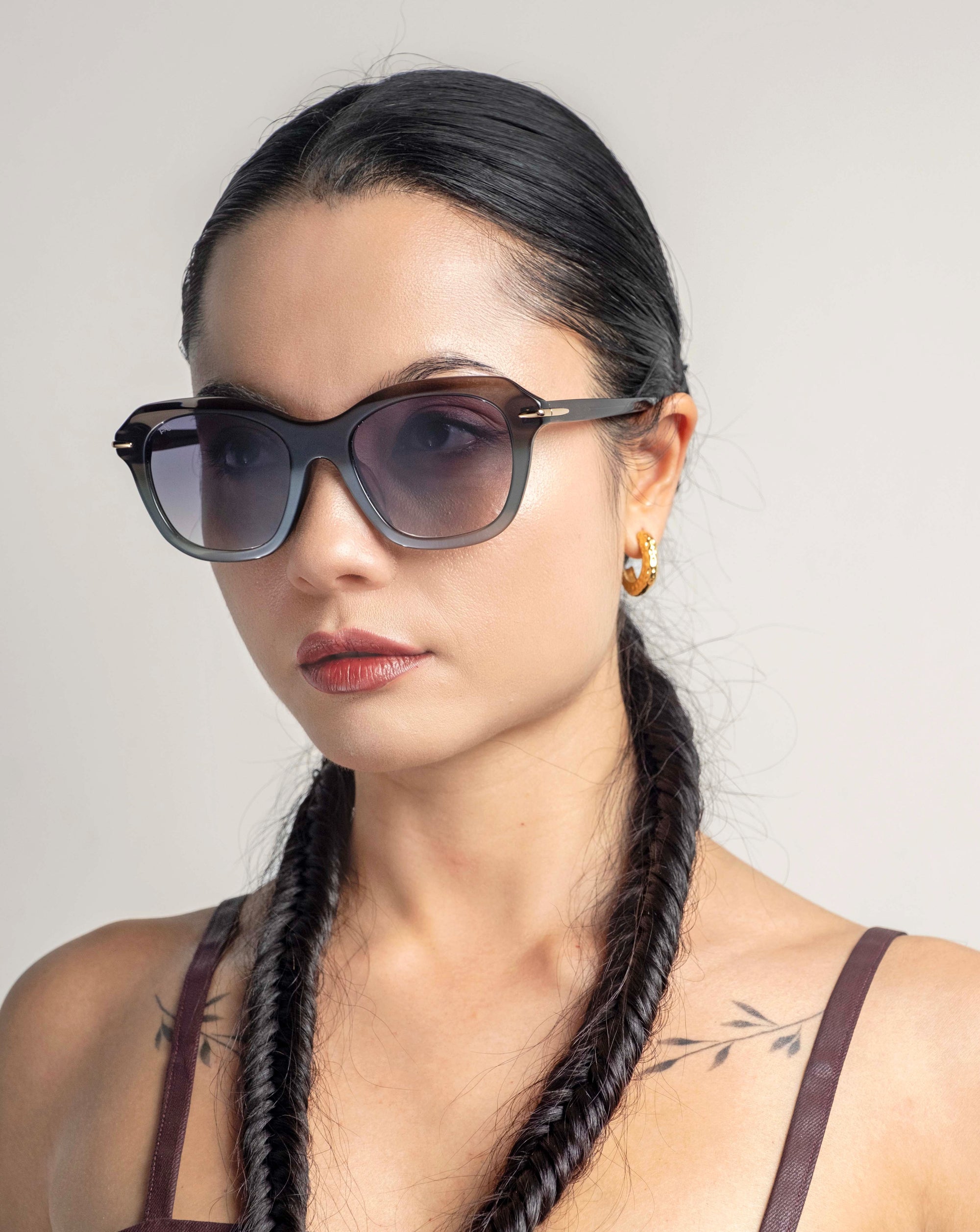 A woman with long dark hair styled in two side braids wears oversized cat-eye Helene sunglasses by For Art's Sake®. She has a neutral facial expression, minimal makeup, and small hoop earrings. Her shoulder tattoos are partially visible. The background is plain and light-colored.