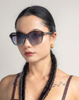 A woman with long dark hair styled in two side braids wears oversized cat-eye Helene sunglasses by For Art's Sake®. She has a neutral facial expression, minimal makeup, and small hoop earrings. Her shoulder tattoos are partially visible. The background is plain and light-colored.