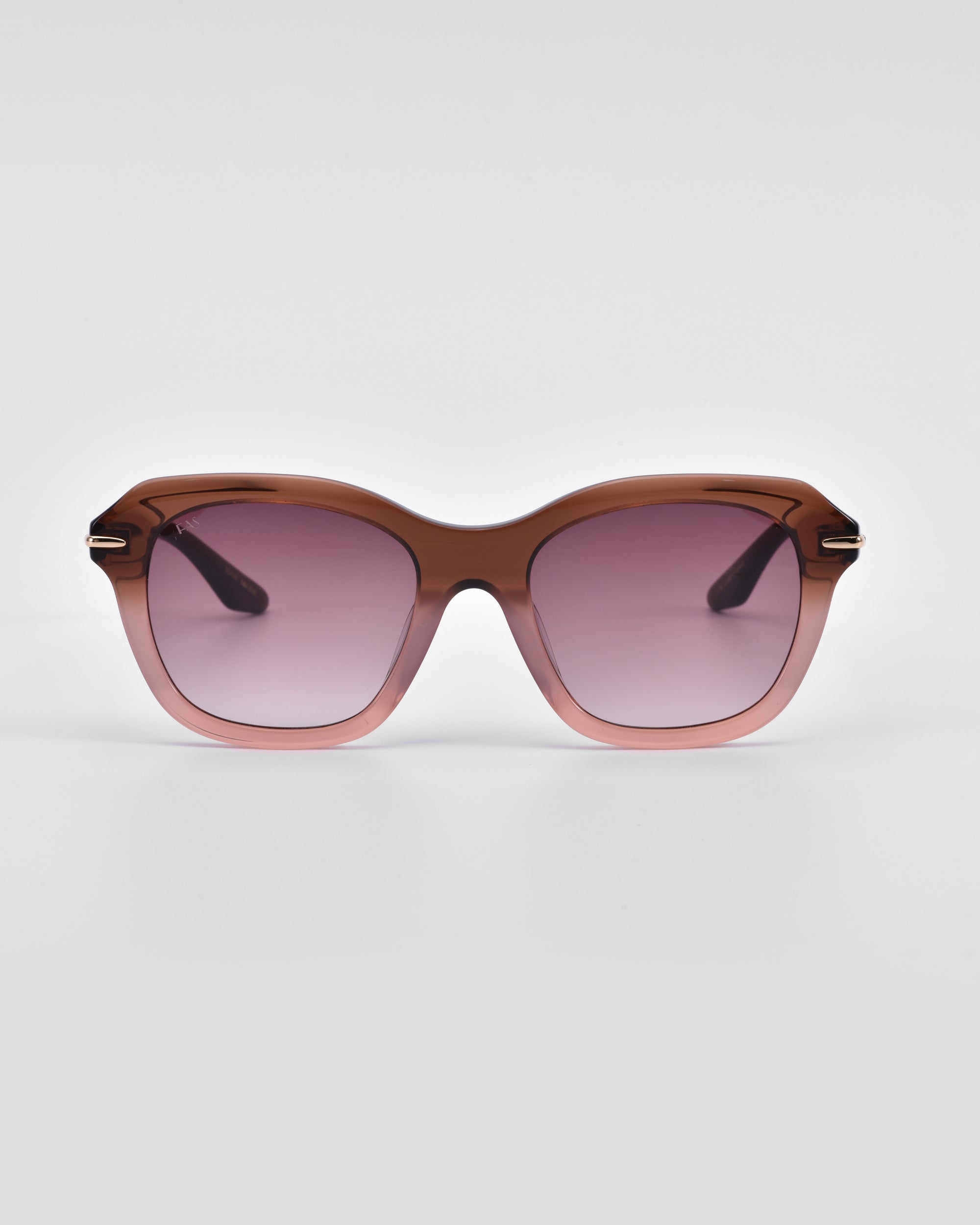 A pair of stylish, oversized cat-eye sunglasses with a gradient brown-pink frame and pink-tinted lenses. The arms of the glasses are black and slightly curved, featuring metallic accents near the hinges. The Helene by For Art's Sake® stands out against a plain white background.