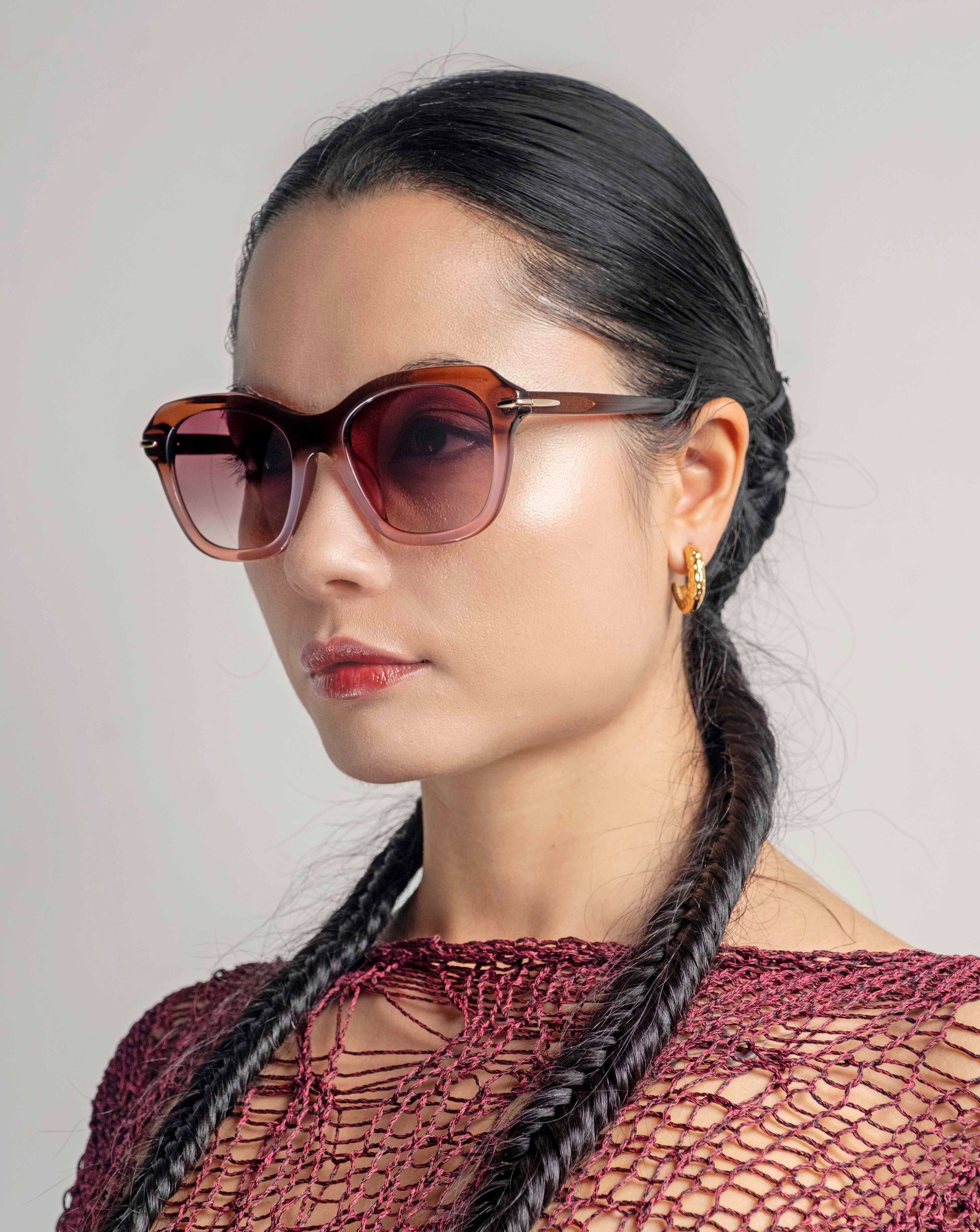 A woman with long, black braids wears oversized cat-eye sunglasses "Helene" by For Art's Sake®. She has gold hoop earrings and a calm expression, facing slightly to the right. The background is a plain, neutral tone.