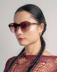 A woman with long, black braids wears oversized cat-eye sunglasses "Helene" by For Art's Sake®. She has gold hoop earrings and a calm expression, facing slightly to the right. The background is a plain, neutral tone.