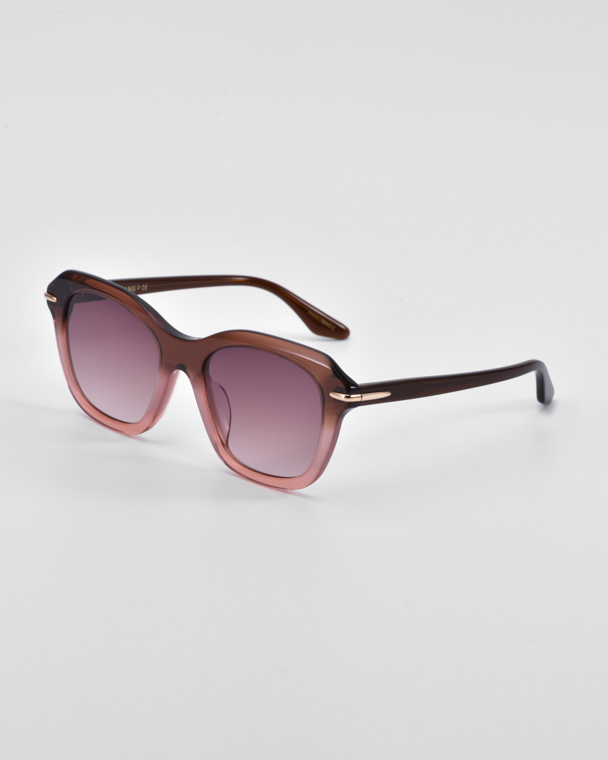 A pair of oversized cat-eye sunglasses with gradient lenses that transition from dark to light pink. The frame is a sleek, dark brown with 18-karat gold plating near the hinges. The design is chic and modern, making it a true statement piece suitable for both casual and formal wear. Introducing "Helene" by For Art's Sake®.