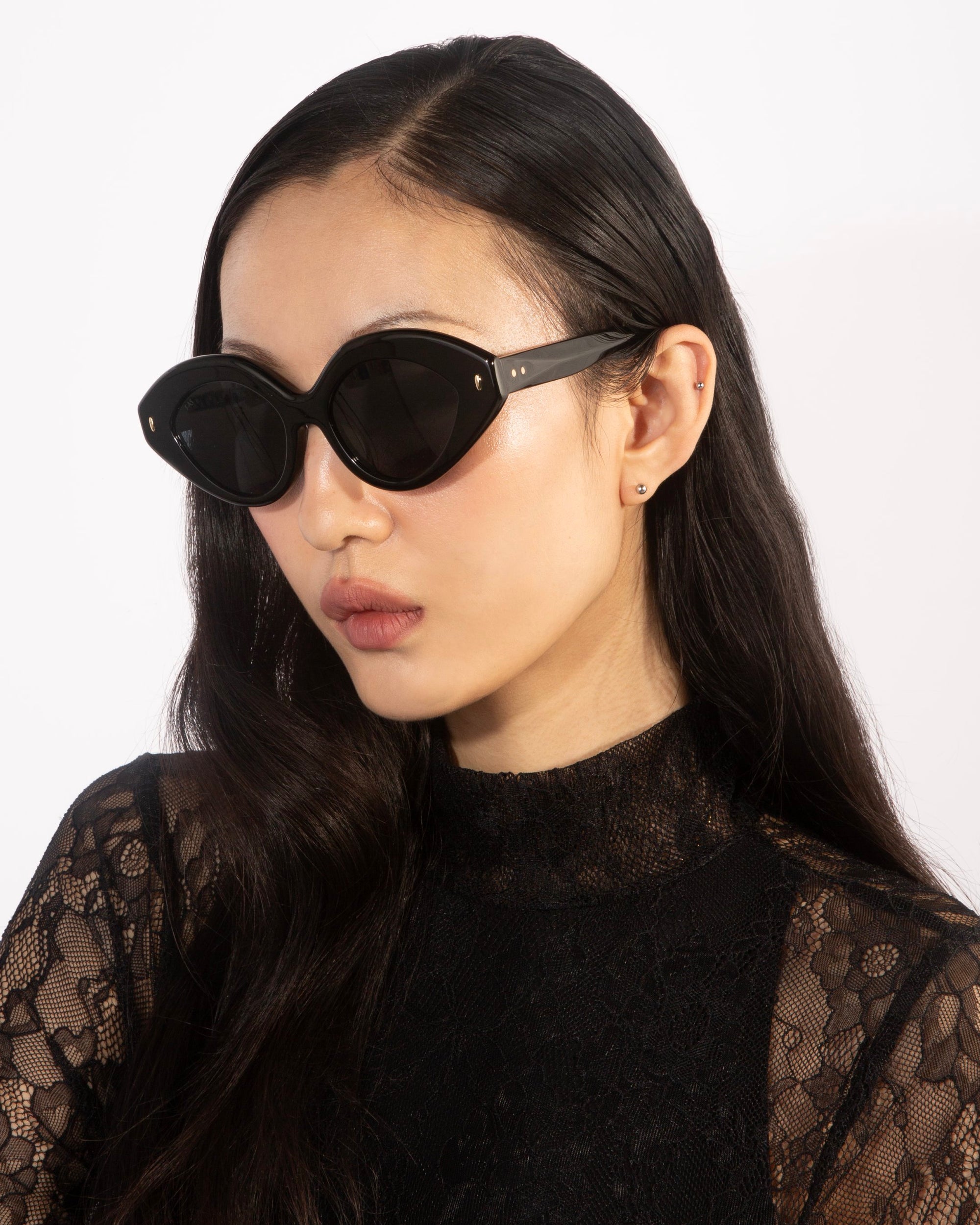 A woman with long, dark hair wears oversized oval Vera sunglasses by For Art&#39;s Sake® and a black lace top. She is facing slightly to the side, showcasing the elegant design of her outfit and accessories against a plain, white background. Her black acetate frames from For Art&#39;s Sake® not only add style but also offer sun protection.