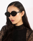 A woman with long, dark hair wears oversized oval Vera sunglasses by For Art's Sake® and a black lace top. She is facing slightly to the side, showcasing the elegant design of her outfit and accessories against a plain, white background. Her black acetate frames from For Art's Sake® not only add style but also offer sun protection.