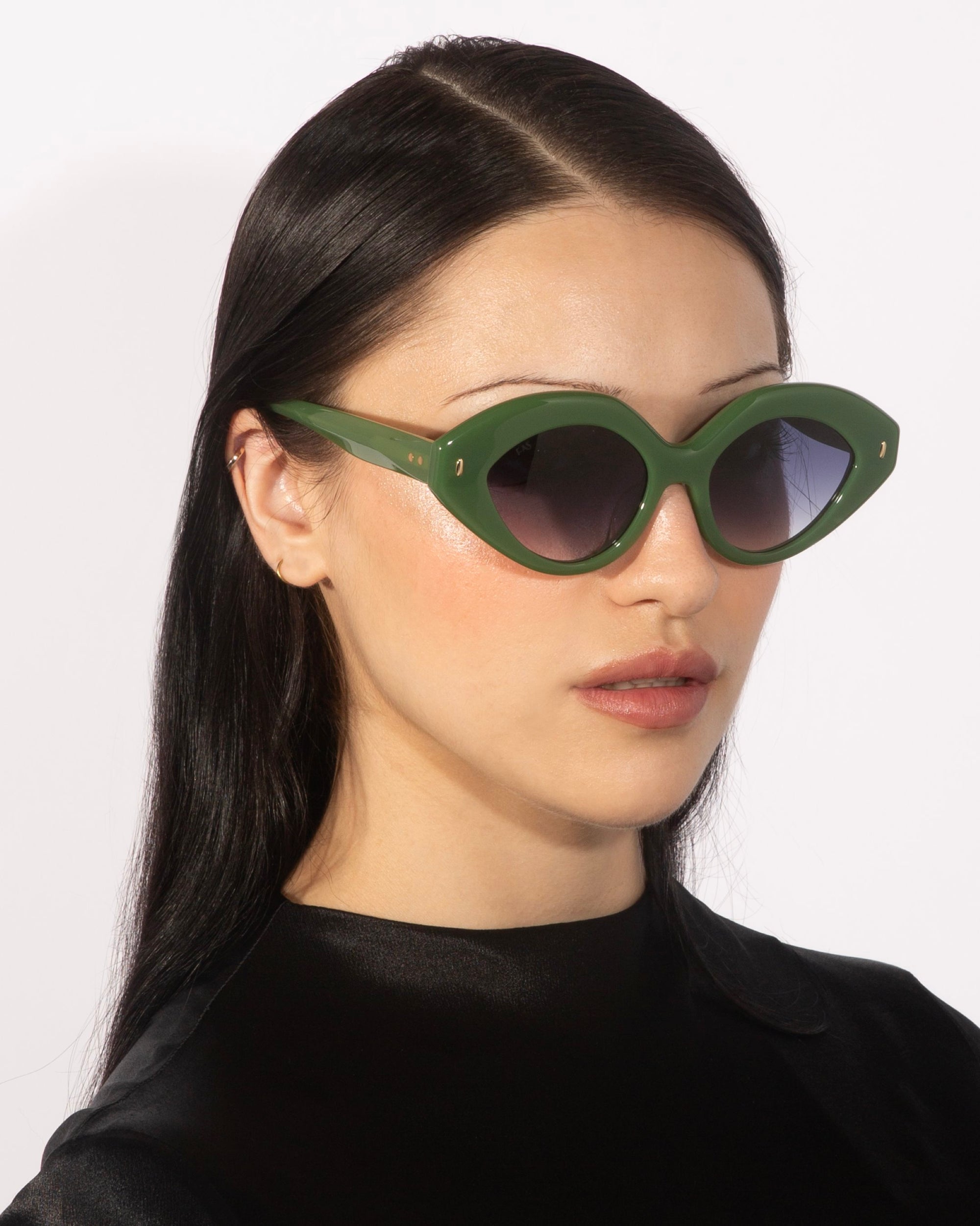 A person with long, dark hair is seen wearing the Vera green cat-eye statement sunglasses with dark lenses from For Art&#39;s Sake®, paired with a black top. The background is plain white.