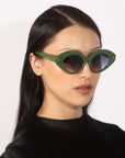 A person with long, dark hair is seen wearing the Vera green cat-eye statement sunglasses with dark lenses from For Art's Sake®, paired with a black top. The background is plain white.