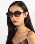 A woman with long, dark hair is wearing the oversized, brown-tinted Vera sunglasses with black acetate frames by For Art's Sake®. She is dressed in a black lace-embellished top and is looking slightly to her left. The plain white background showcases her minimalist and chic style.