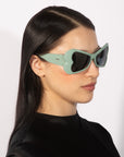 A person with long, straight black hair is adorned in oversized, light green "Bubblegum" sunglasses by For Art's Sake®, featuring a unique, wavy design. They wear a black top and face to the right with a neutral expression, set against a plain white background.