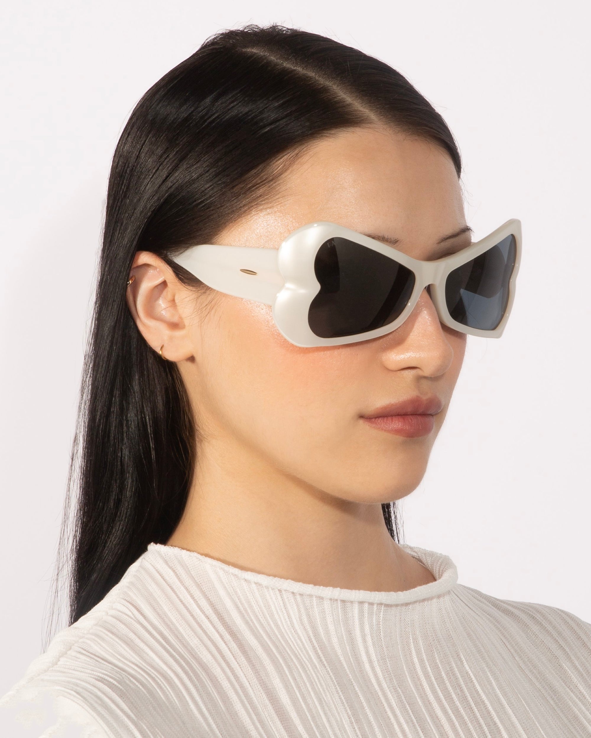 A person with long, straight, dark hair sports the bold Bubblegum white heart-shaped sunglasses by For Art&#39;s Sake®, paired with a textured white top. The plain white background accentuates the stylish look. Their expression is neutral as they face slightly to the side.