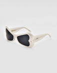 A pair of stylish For Art's Sake® Bubblegum sunglasses features white frames with heart-shaped lenses tinted in dark black. This bold design is both playful and chic, creating a striking fashion statement. Made from sustainable materials, the sunglasses are positioned against a neutral, light-colored background.