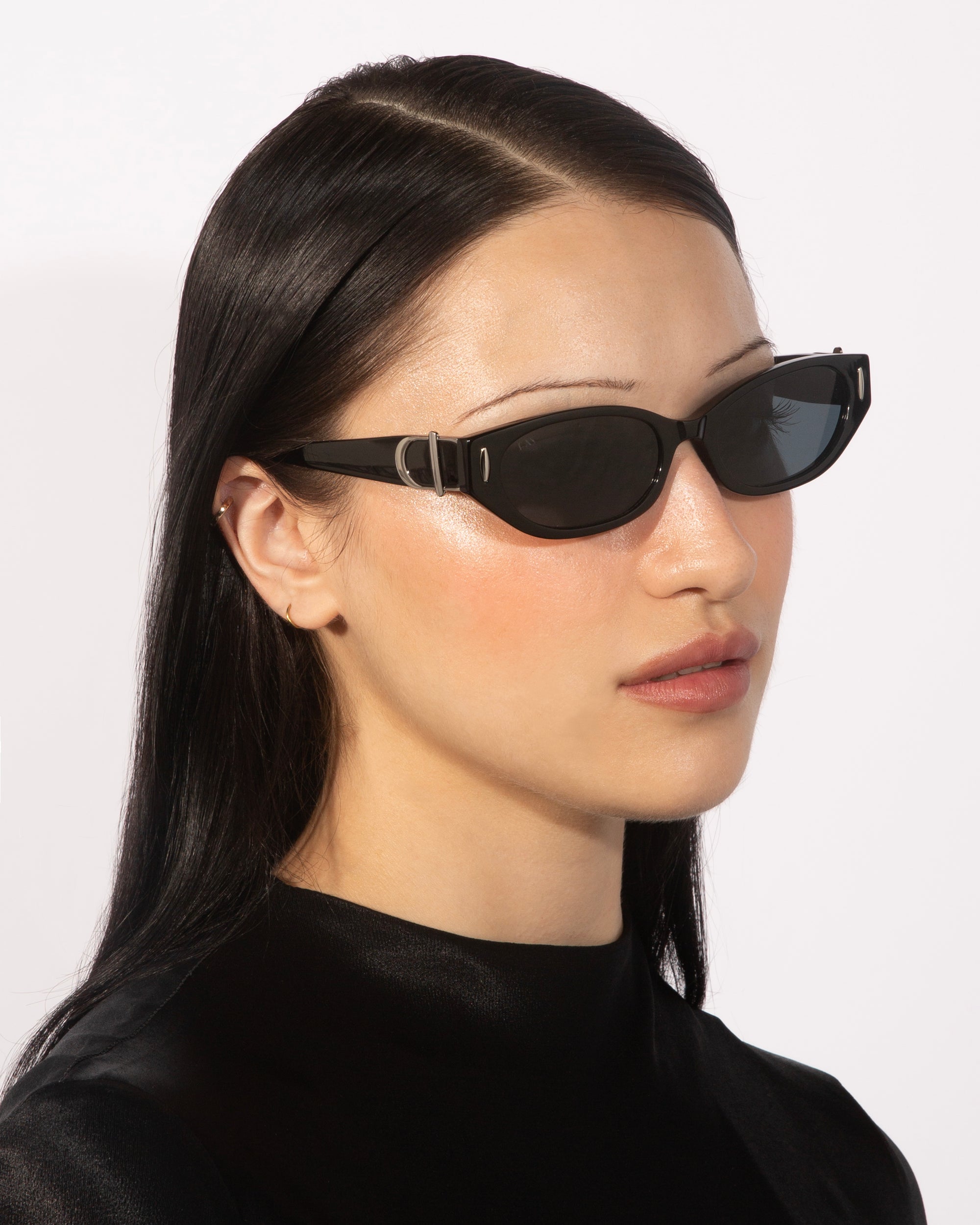 A person with long, straight black hair is wearing sleek, black rectangular sunglasses with narrow frames that exude 90s chic. They are dressed in a black top against a plain white background. Their For Art&#39;s Sake® Skinny Latte sunglasses showcase polished acetate, and they gaze slightly to the right with a calm expression.