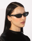 A person with long, straight black hair is wearing sleek, black rectangular sunglasses with narrow frames that exude 90s chic. They are dressed in a black top against a plain white background. Their For Art's Sake® Skinny Latte sunglasses showcase polished acetate, and they gaze slightly to the right with a calm expression.