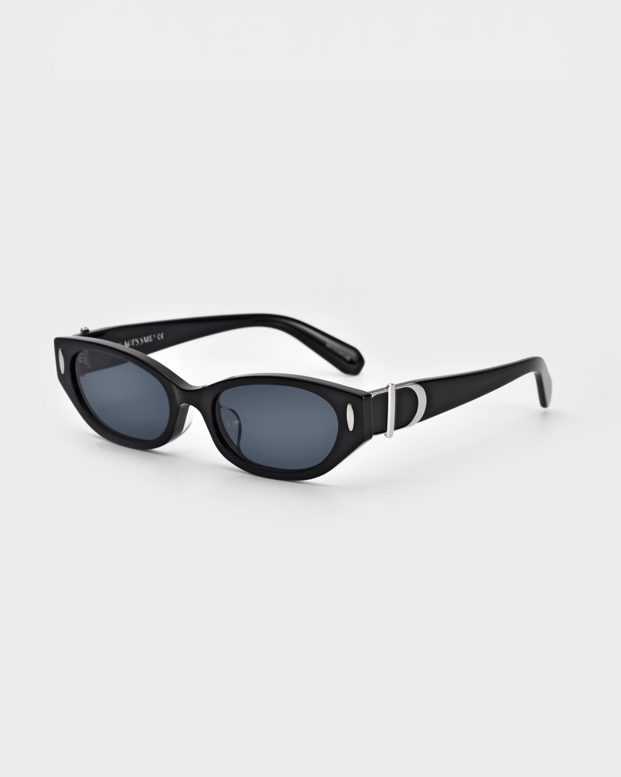 A pair of stylish For Art&#39;s Sake® Skinny Latte sunglasses featuring black-frame, oval dark-tinted lenses, and sleek arms. The right arm is adorned with a small metallic &quot;D&quot; ornament near the hinge, reflecting a minimalist design. The background is plain white.