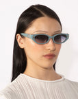 A person with long black hair wears For Art's Sake® Skinny Latte sunglasses and a white ribbed top. The background is plain white.